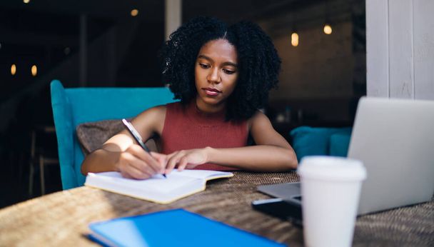 Dark skinned hipster girl writing information in knowledge notepad while sitting at table with modern technology for research and e learning, African American female student preparing to exam - Zdjęcie, obraz