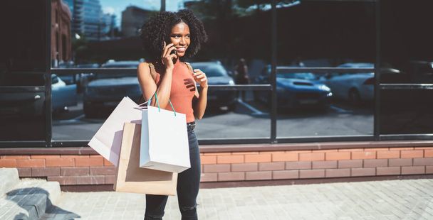 Happy black female teenager with paper bags calling to friend for discussing Cyber Monday discounts using roaming connection on cellphone, positive woman in casual wear smiling from funny joke - Foto, Imagem