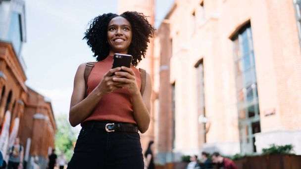 Below view of positive hipster girl looking away and smiling during online communication with followers from social networks, African American female teenager with modern technology in hand - Foto, immagini