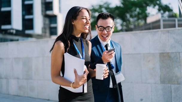 Diverse stylish man and woman with id badges having coffee and walking down street while sharing smartphone and laughing in sunshine - Photo, image