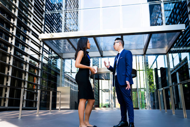 Side view of African American woman and Asian man in formal clothes standing against contemporary office building having coffee break and talking  - Photo, image