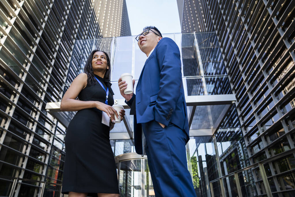 From below of confident multiracial woman and man with coffee cups standing on background of modern city building smiling away - Foto, imagen