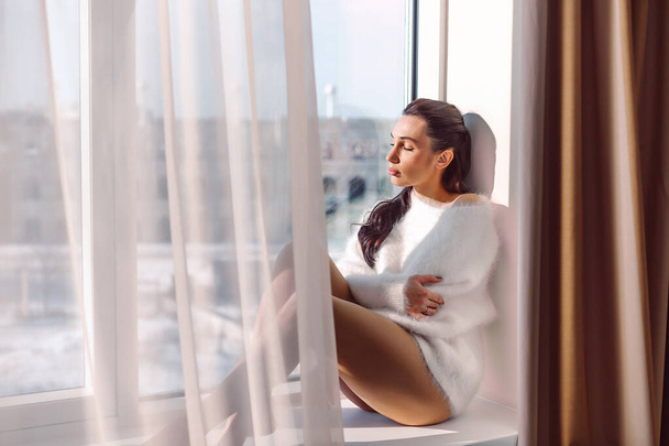 Beautiful brunette girl in a white sweater and white lingerie sits on a windowsill near the window, the sun burns her, she looks pleased - Фото, зображення