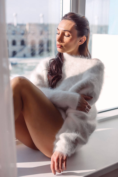 Krásná brunetka dívka v bílém svetru a bílé spodní prádlo sedí na parapetu u okna, slunce ji pálí, vypadá potěšeně - Fotografie, Obrázek