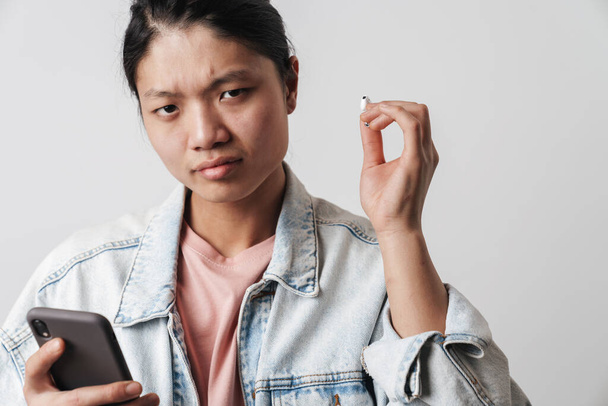Image of displeased asian man using cellphone and wireless earphone isolated over white background - Foto, immagini