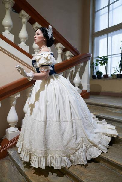 A beautiful girl, in a romantic dress with a low neck, retro style, with dark hair decorated with a white feather.In full growth, goes down the stairs. Historical reconstruction                                   - Photo, Image