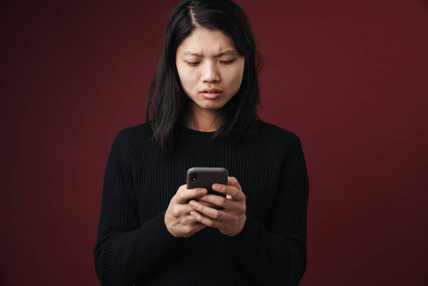 Image of perplexed asian man posing and using cellphone isolated over burgundy background - Fotografie, Obrázek