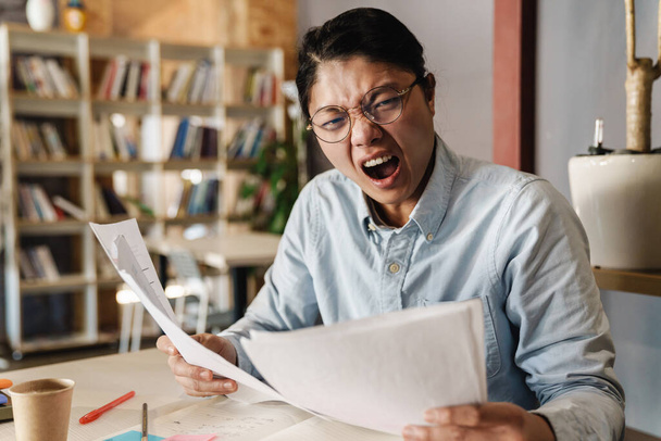 Image of irritated asian man in eyeglasses expressing shock while doing homework and sitting at table - Photo, image