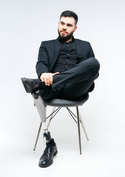 Behinderter junger Mann mit Beinprothese sitzt auf einem Stuhl im Studio vor weißem Hintergrund, Konzept der künstlichen Gliedmaßen - Foto, Bild