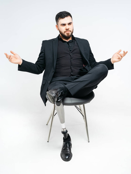 Niepełnosprawny młody człowiek z protezą nogi siedzący na krześle w studio na białym tle, sztuczna koncept kończyny - Zdjęcie, obraz