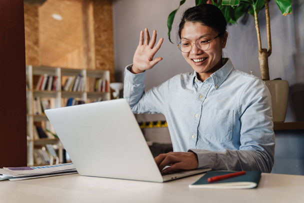 Image of joyful handsome asian man in eyeglasses using laptop and waving hand while sitting at table - Foto, afbeelding
