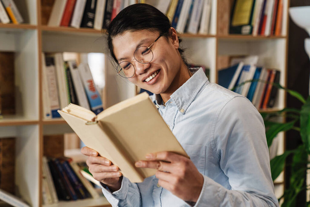 Afbeelding van de vrolijke knappe Aziatische man in een bril glimlachend en leesboek terwijl hij in de bibliotheek staat - Foto, afbeelding