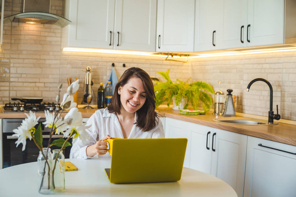 Kobieta pijąca herbatę z żółtego kubka pracująca na laptopie. kuchnia w tle. Koncepcja wolnego strzelca - Zdjęcie, obraz