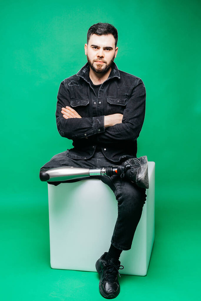 Fogyatékkal élő fiatalember műlábbal ül a stúdióban a zöld háttér felett, mesterséges végtag koncepció - Fotó, kép