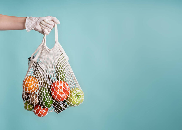 Zero waste concept. Coronavirus food delivery. Woman in glove holding a white mesh bag with vegetables isolated on blue background with copy space - Photo, Image