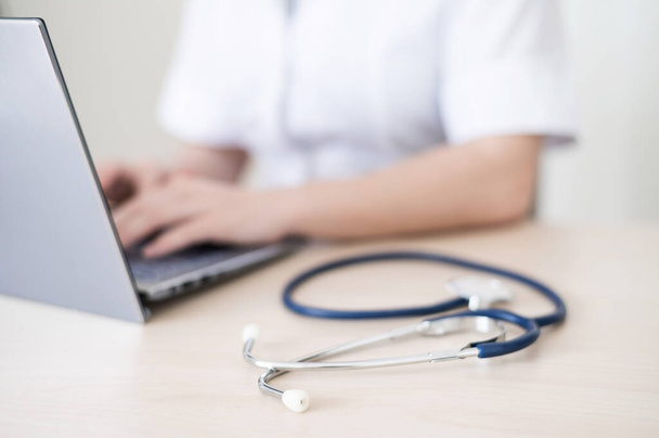 Az egyenruhás, felismerhetetlen női orvos laptopot használ. Az arctalan orvos jelentést ír az íróasztal számítógépéről. Vágott. - Fotó, kép
