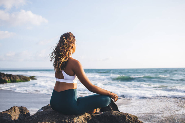 Yoga on beautiful Indian beach performed by attractive young African American girl dressed in sport tracksuit, evening spiritual training near ocean by calm female in lotus pose meditating - Photo, Image
