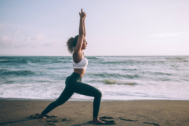 Attractive young woman in casual tracksuit doing virabhadrasana one exercise during outdoors training near sea, female enjoying yoga healthy lifestyle for relaxing minds and get soul harmony - Foto, Imagem