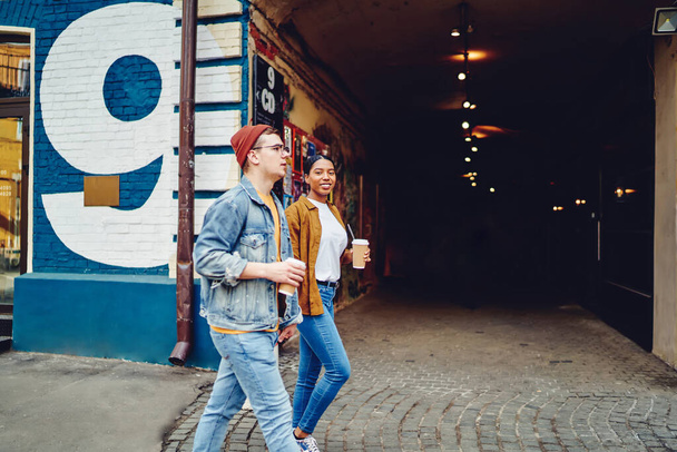 Stylish casual dressed multicultural young man and woman enjoying tasty coffee beverage and walk in modern city on weekend.African american hipster girl with caucasian friend strolling on street - Фото, зображення