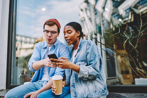African american young woman showing app on smartphone wondered hipster guy dressed in casual denim wear sitting outdoors with coffee.Multicultural friends watching video on website on cellular - Photo, Image