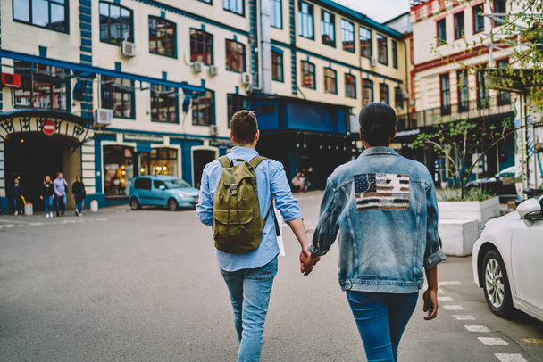 Back view of stylish casual dressed couple in love holding hands each other and strolling on street of cozy city during common trip on weekend.Diverse girlfriend and boyfriend walking together in town - Фото, зображення