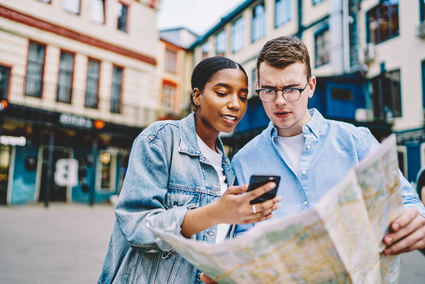 Pensive caucasian young man holding travel map and looking for route together with african american girlfriend which showing smartphone with online navigator in app standing in urban setting of city - Valokuva, kuva