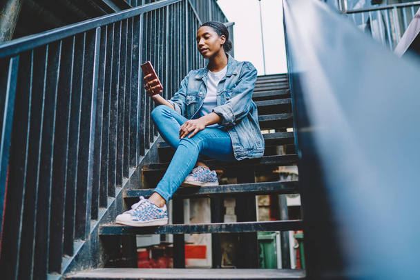 Afrikai amerikai fiatal nő blogger gazdaság smartphone eszköz és üzenetküldés az online chat követők segítségével 4G internet kapcsolat.Sötét bőrű hipster lány farmerben viselni olvasási hírek mobil - Fotó, kép