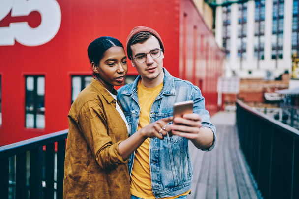 Multiracial couple in love dressed in trendy wear checking new app for searching places of interest in city on cellphone, male and female millennial hipsters read news from social network on smartphon - Φωτογραφία, εικόνα