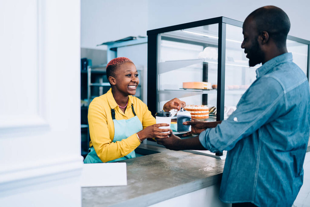 Adult black female in apron smiling and giving takeaway hot beverage and dessert to African American man while working in cozy cafe - Photo, Image