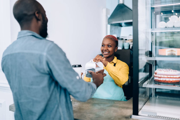 Friendly African American woman in apron friendly smiling and giving hot beverage and food to anonymous male customer while working in modern cafe - Foto, Imagen
