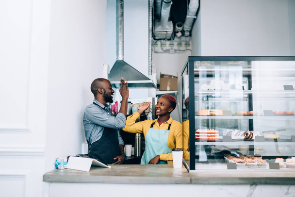 African American man and woman in aprons smiling and giving high five to each other while working in modern cafe together - Foto, Imagem