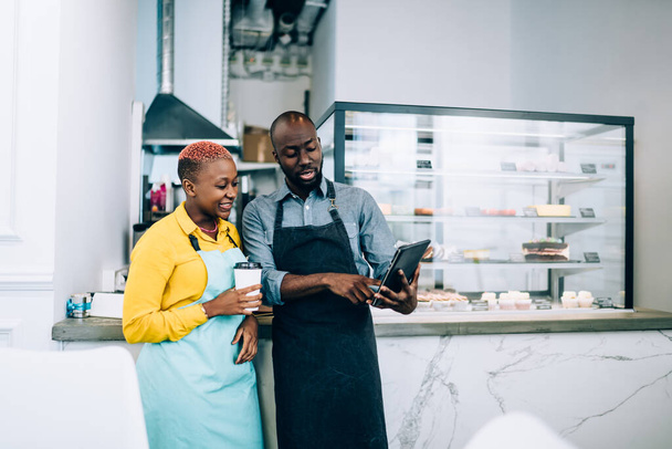 Bearded African American man in apron demonstrating modern tablet to female colleague with cup of hot beverage while working in cafe together - Foto, immagini