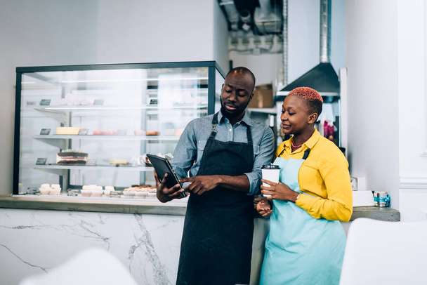 African American man and woman in aprons enjoying hot beverage and using modern tablet while standing near counter during break in cafe - Foto, Bild