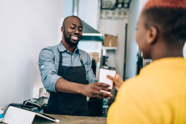 Handsome African American man in apron friendly smiling and giving cup of fresh hot beverage to unrecognizable black woman while working in modern cafe - Fotoğraf, Görsel