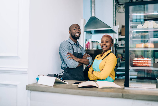 Positive African American man and woman with crossed arms smiling and looking at camera while standing behind counter in cozy bakery - Foto, Bild
