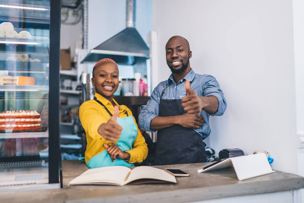 African American man and woman in aprons smiling and showing thumb up gesture while recommending service of small cozy bakery - Φωτογραφία, εικόνα