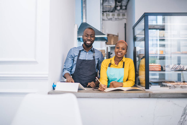 Afroamerykanin mężczyzna i kobieta w fartuchach uśmiechający się i patrzący w kamerę podczas używania tabletu i pisania w dzienniku stojąc za ladą w małej kawiarni - Zdjęcie, obraz