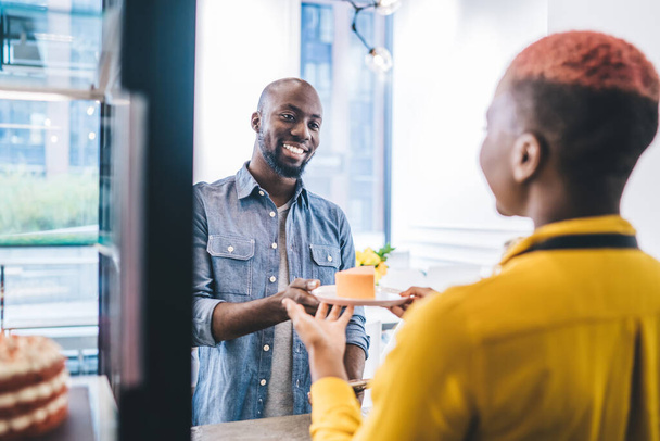 Handsome African American male friendly smiling and taking piece of yummy cake from anonymous female barista while making order in small cafe - Photo, Image