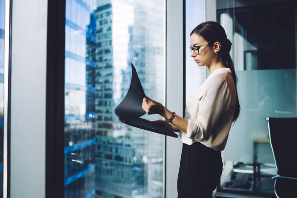 Side view of beautiful elegant woman in glasses reading documents in file while standing alone in contemporary office near window - Fotografie, Obrázek
