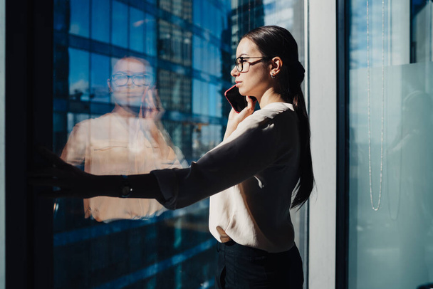 Side view of elegant modern woman in glasses standing near office window in sunlight having call on mobile phone and looking away - Foto, imagen