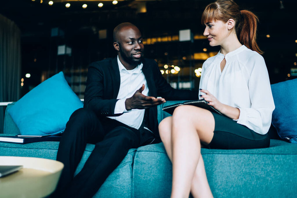 Handsome black businessman and young formal woman with tablet sitting on sofa in cafe and chatting while having professional business meeting  - Foto, Bild