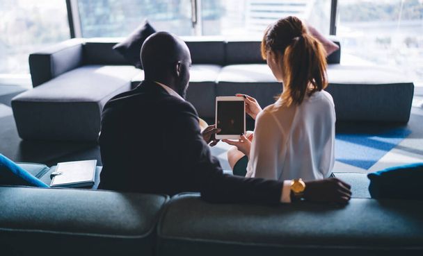 Back view of contemporary elegant black man and formal woman sitting on couch in hallway and sharing wireless tablet coworking together - Foto, Imagem