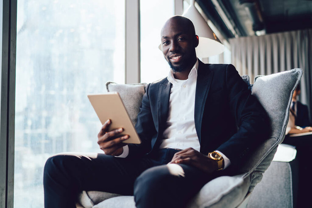 Handsome confident black man in suit holding tablet while sitting in armchair and smiling at camera in hallway of office - Zdjęcie, obraz