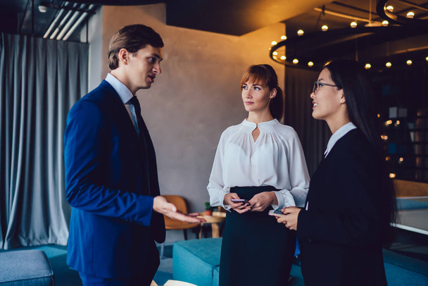 Side view of elegant man with young female colleagues standing in stylish office hallway and talking friendly looking at each other - Фото, зображення