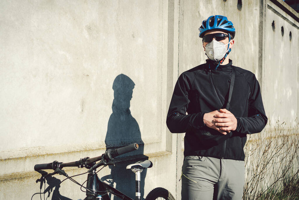 Koruyucu yüz maskesi takmış bir bisikletçi gri beton duvarın arka planında bir bisikletin yanında duruyor. Solunum cihazında tanımlanamayan kurye. Bir salgın sırasında teslimat servisi. - Fotoğraf, Görsel