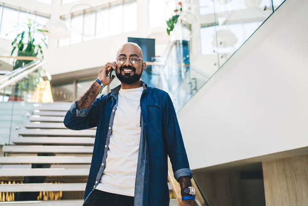 Cheerful handsome ethnic man with beard standing on stairway in light hallway smiling at camera while having call on mobile phone - Фото, изображение