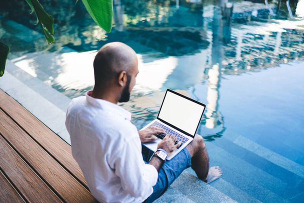 From above of man in shirt sitting on edge of pool with legs in water while using laptop with white empty screen - Zdjęcie, obraz