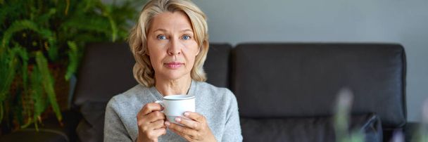 Deliciosa bebida.Mujer mayor sentada en el sofá de su sala de estar y sosteniendo una taza de café
. - Foto, Imagen