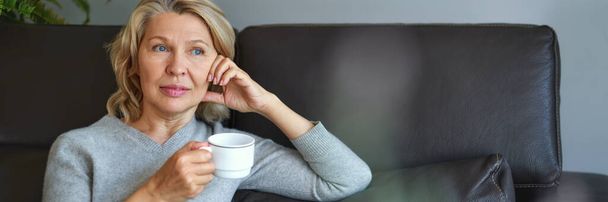 Літня жінка сидить на дивані у вітальні і тримає чашку кави
. - Фото, зображення