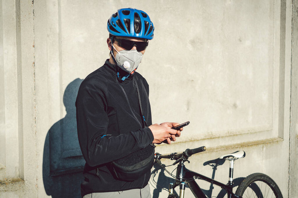 Der Kurier mit Schutzmaske auf dem Fahrrad hielt an und beobachtete die Telefonanwendung. Lieferservice in Quarantäne. Ein Mann mit Atemschutzmaske benutzt ein Mobiltelefon. Fahrradkurier funktioniert. - Foto, Bild